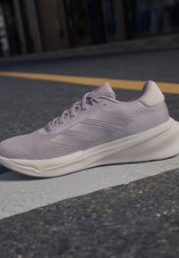 Zapatilla de running de color púrpura claro con parte superior de malla, que presenta tres rayas de acento, una suela acolchada y una textura suave.