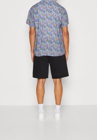 Korte mouwen, gekraagde blouse met een meerkleurig bloemenpatroon; gecombineerd met zwarte shorts en witte sneakers; zichtbare sokken.