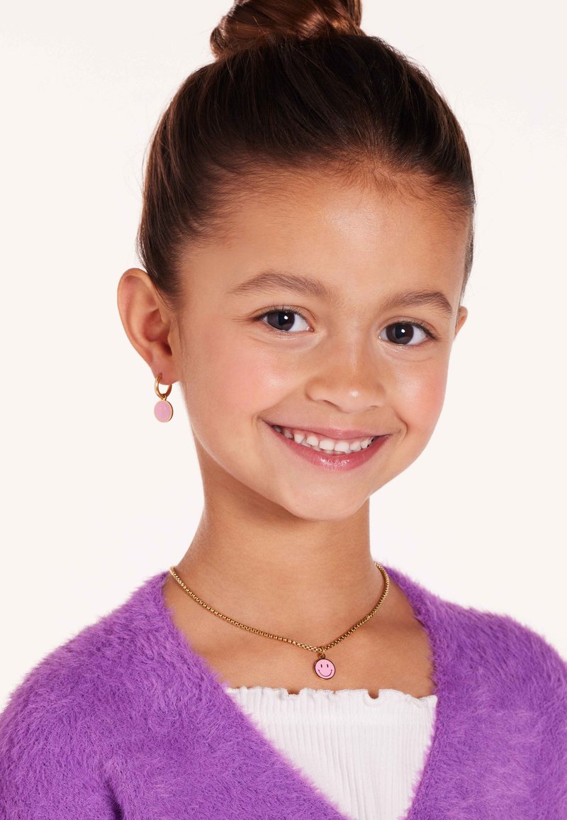 Gouden ketting met een roze smiley-hanger, aangevuld met bijpassende roze creolen. Draagt het met een fluffy paarse trui.
