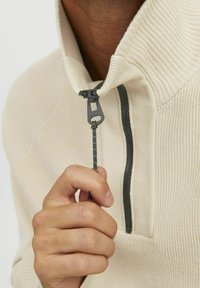 Beige pullover met een hoge kraag en een halve rits. Heeft een geribbelde textuur, zwarte rits en koord voor een verstelbare pasvorm.