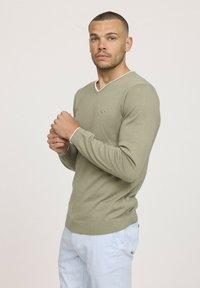 Pull vert kaki avec un col en V et des bordures blanches contrastantes au niveau du col et des poignets. Design ajusté, fabriqué en tricot doux.