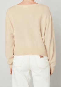 Béžový pletený svetr s volným střihem, širokými rukávy a žebrovaným lemem, doplněný bílými džínami s vysokým pasem a červenou značkovou cedulkou.