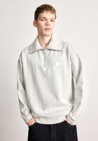 Ανοιχτό γκρι μπλουζάκι με ψηλό γιακά, κλείσιμο με μισό φερμουάρ και μικρό λογότυπο. Κατασκευασμένο από απαλό, υφασμάτινο ύφασμα με μακριά μανίκια.