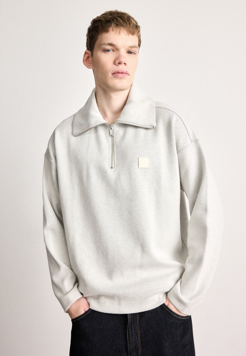 Ανοιχτό γκρι μπλουζάκι με ψηλό γιακά, κλείσιμο με μισό φερμουάρ και μικρό λογότυπο. Κατασκευασμένο από απαλό, υφασμάτινο ύφασμα με μακριά μανίκια.