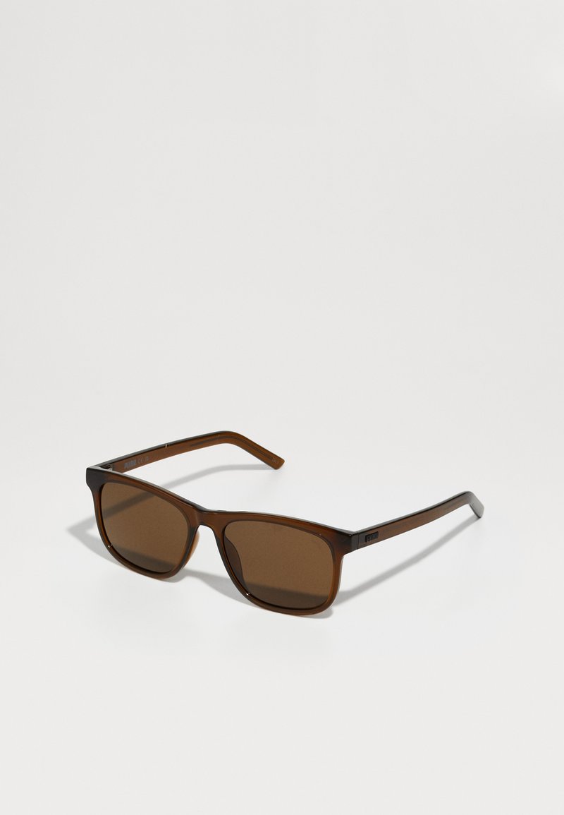 Óculos de sol retangulares castanhos com lentes castanhas escuras. Armação de plástico transparente, hastes retas e uma marcação subtil na têmpora.
