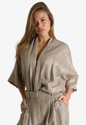 Donna che indossa una camicetta a righe metallizzate beige con maniche larghe e pantaloni a vita alta abbinati con le mani nelle tasche.