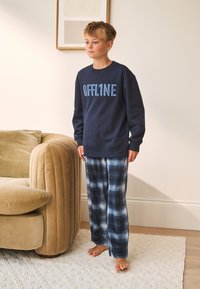 Tengerészkék pulóver "OFFLINE" felirattal, kék kockás flanel pizsamanadrággal párosítva. Világos textúrájú szőnyegen áll, egy ívelt kanapé mellett.