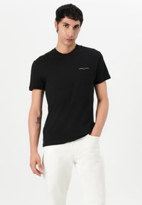 Fekete pamut póló rövid ujjakkal, kerek nyakkal és egy kis fehér "TOMMY JEANS" logóval a bal mellkason. Fehér nadrággal párosítva.