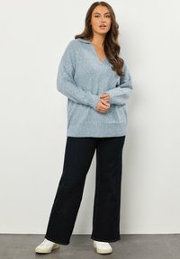 Helles Blau, oversized, gestrickter Pullover mit V-Ausschnitt, geripptem Saum und langen Ärmeln, kombiniert mit weit geschnittenen, dunklen Jeans und weißen Sneakers.