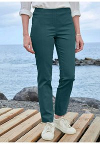 Donkerblauwe skinny broek met een hoge taille, gladde textuur, voornaaddetail, gecombineerd met witte sneakers, geplaatst op houten planken nabij water.