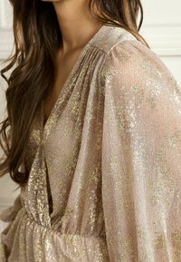 Robe transparente et scintillante en doux rose pâle avec un éclat doré. Elle présente un design drapé à encolure en V, des manches fluides, un tissu texturé et une taille élastique.