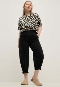 Donna che indossa una maglietta a maniche corte con stampa leopardata, pantaloni neri a vita alta abbottonati e scarpe beige in pelliccia slip-on, in piedi con una mano in tasca.