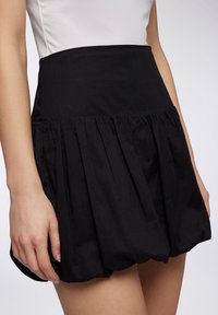 Gonna plissettata nera con vita alta, realizzata in tessuto leggero. Presenta un design arricciato e una superficie liscia. Nessuna parte metallica visibile.