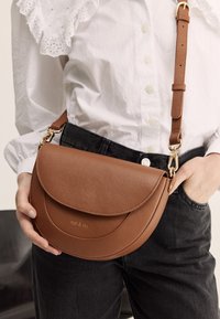 Brown leather crossbody bag with a curved flap closure, textured surface, gold hardware accents, and an adjustable strap.