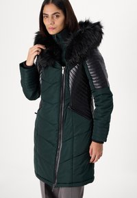 Padded coat in deep green with black faux fur collar. Features quilted, textured sleeves and a front zipper with side pockets.