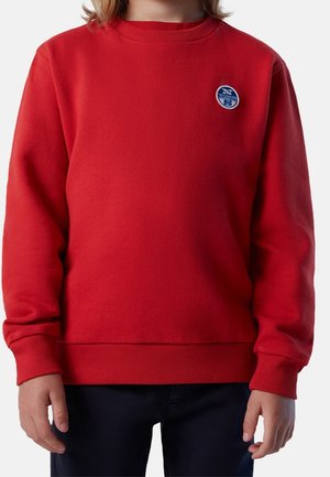 Enfant portant un pull rouge à manches longues avec un petit logo rond North Sails sur la poitrine et un pantalon foncé.