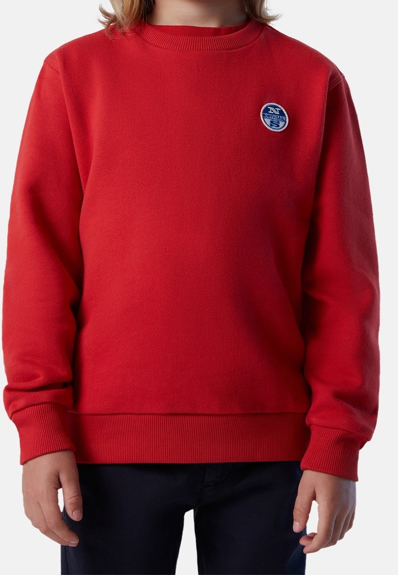 Enfant portant un pull rouge à manches longues avec un petit logo rond North Sails sur la poitrine et un pantalon foncé.