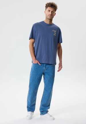 Hombre de pie con una mano en el bolsillo, vestido con camiseta azul con la frase "c’est la vie", jeans azul claro y zapatillas blancas sobre fondo blanco.