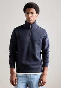Felpa con zip di colore blu navy e colletto alto, con un dettaglio di cucitura sul petto e polsini a costine. Tessuto morbido e testurizzato.