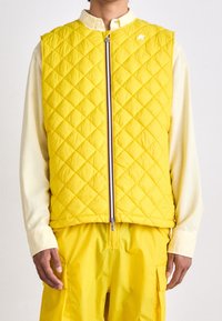Persona che indossa un gilet trapuntato giallo sopra una maglietta a maniche lunghe color crema, con pantaloni gialli coordinati, in piedi su uno sfondo bianco.