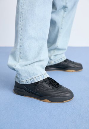 Personne debout sur un tapis bleu portant un jean bleu clair et des chaussures de sport noires avec des semelles en gomme.