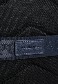 Musta teksturoitu kangas timantinmuotoisella kuviolla, jossa on laivastonsininen logoplässi, jossa lukee "U.S. POLO ASSN", ja sileä teksturoitu hihna.