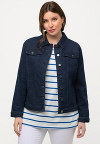 Giacca in denim scuro con chiusura a bottoni, due tasche sul petto e colletto strutturato, abbinata a una camicia bianca e blu a righe sotto.