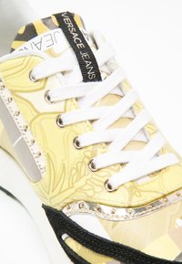 Gros plan sur une sneaker Versace Jeans jaune à motifs avec des lacets blancs, des œillets argentés, des accents noirs et des détails à clous dorés.