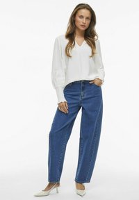 Balta gara piedurkņu blūze ar V-veida kakla izgriezumu un strukturētu audumu, kombinēta ar augsto vidukļa zilām taisnām biksēm un baltām, smailām kurpēm.