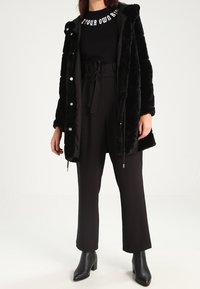 Černý kabát z umělé kožešiny s kapucí, předními patentkami a bočními kapsami. Nesený přes černý top a černé kalhoty s vysokým pasem.