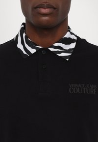 Black polo shirt featuring a black and white zebra print collar, with a textured fabric, two buttons, and 'VERSACE JEANS COUTURE' logo in grey.