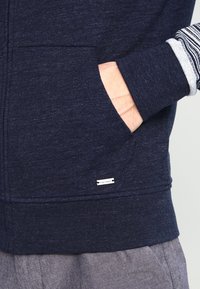 Marineblå zip-up hoodie med en strukturerad yta, med randig insida, sidofickor och en diskret metallisk logotagg nära nederkanten.