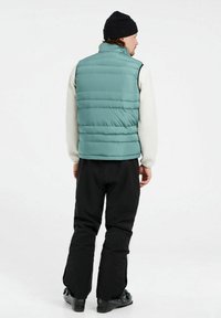 Jasnoniebieska kamizelka puchowa z wysokim kołnierzem, pionowymi szwami i gładką fakturą, noszona na białej koszuli z długim rękawem i czarnych spodniach.