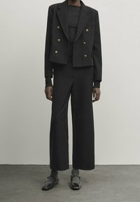 Musta räätälöity kaksirivinen bleiseri kultaisilla nappeilla, yhdistetty leveälahkeisiin housuihin ja mustiin kenkiin, teksturoitu kangas, tyylikäs muotoilu.