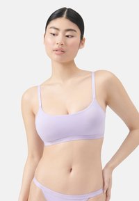 Eine Frau mit glattem schwarzem Haar trägt einen nahtlosen Lavendel-BH und passende Unterwäsche und steht vor einem schlichten hellen Hintergrund.