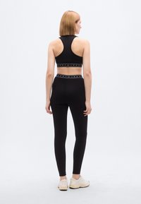 Σετ μαύρου αθλητικού σουτιέν και κολάν, με εφαρμοστή γραμμή, ελαστική μέση με λογότυπο και λεία υφή. Η μοντέλο φοράει αθλητικά παπούτσια.