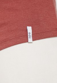 Nahaufnahme eines rostfarbenen Stoffes mit einem weißen "edc"-Markenschild, das in die Naht auf einem hellen strukturierten Hintergrund genäht ist.