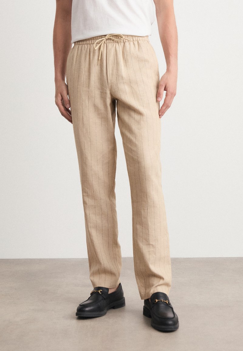 Beige stripete bukser med elastisk linning og justerbar snor. Stoffet ser lett ut med en glatt tekstur. Nøytral passform.