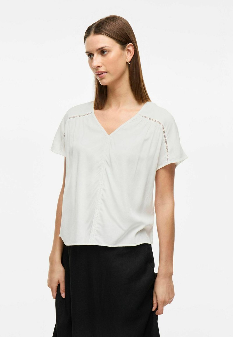Top bianco a maniche corte con scollatura a V, plissettature frontali e cuciture decorative sulle spalle. Tessuto morbido e fitting rilassato.