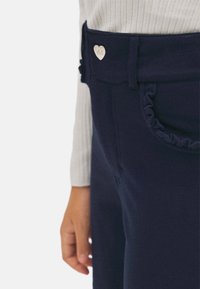 Pantalones azul marino con un detalle de bolsillo con volantes y un botón metálico en forma de corazón plateado en la cintura, hechos de una tela suave.