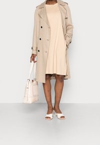 Béžový trenčkot s černými knoflíky přes světle béžové šaty. Bílé lodičky na vysokém podpatku a béžová kabelka přes rameno s přední kapsou.