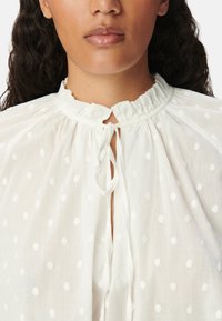 Camicia bianca in tessuto leggero, con colletto a volant, scollatura annodata e punti bianchi a rilievo su tutto il capo.