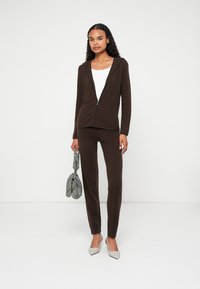 Smeđa zip-up dukserica i koordinirane hlače od mekog materijala, nosi se s bijelim topom i svijetlosivim cipelama s oštrim vrhom. Drži torbu s teksturom.