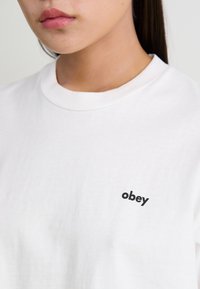 Hvit bomulls t-skjorte med rund hals og en liten svart "obey" logo brodert på venstre side. Glatt tekstur, avslappet design.