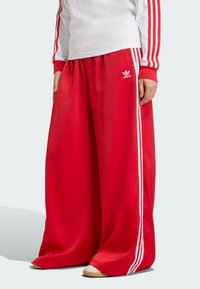 Red wide-leg sweatpants made of synthetic material, featuring white side stripes and an embroidered logo on the left hip.