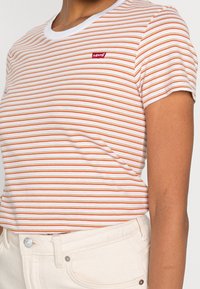 T-shirt rayé orange et blanc, avec un col rond blanc et un petit logo rouge Levi's sur la poitrine. Manches courtes, design ajusté.