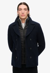 Abrigo de marina de lana con grandes solapas, diseño de doble botonadura y chaqueta de abrigo negra debajo. Líneas limpias y textura suave.