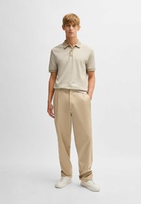 Polo beige à manches courtes, avec deux boutons, associé à un pantalon beige clair et des baskets blanches. Tissu lisse ; coupe décontractée.