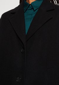 Manteau en laine noir avec de larges revers et deux boutons noirs, porté sur une chemise à col bleu sarcelle.