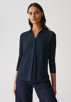 Blusa blu navy scuro con una texture morbida e liscia, un'apertura frontale e maniche a tre quarti. Abbinata a jeans scuri per un contrasto.